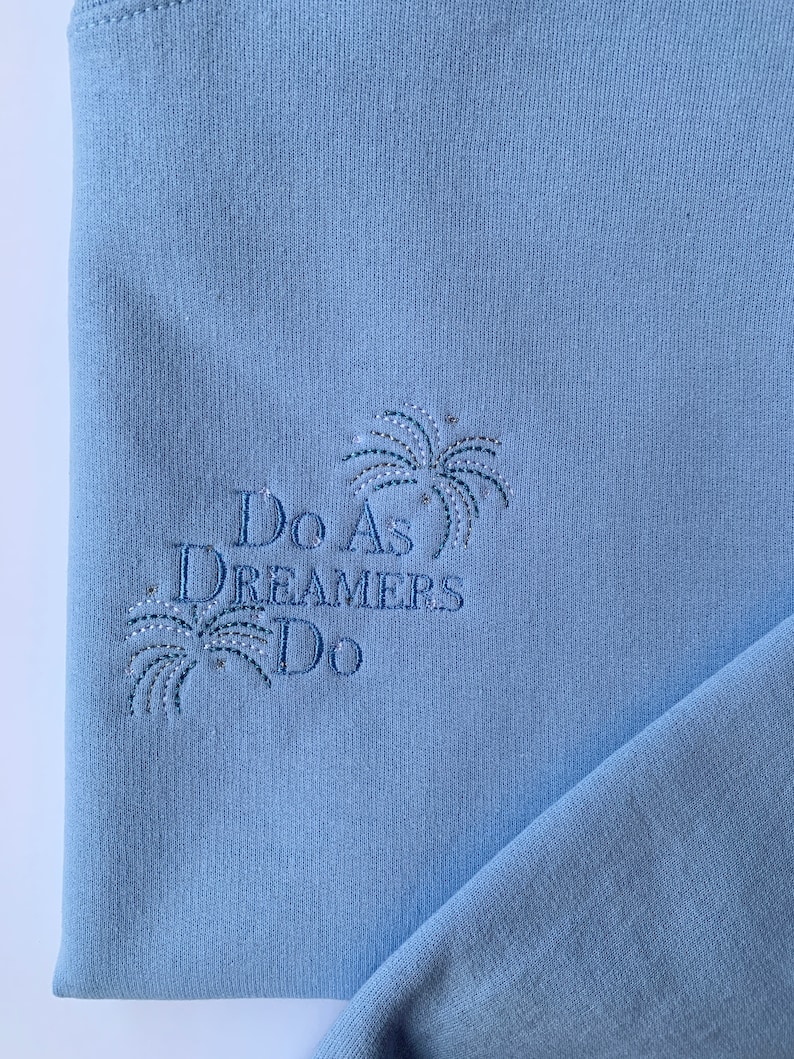 Op de afbeelding: Lichtblauwe trui met de tekst "Do As Dreamers Do" geborduurd in donkerblauwe draad. De tekst wordt omringd door een gestileerd vuurwerkontwerp.