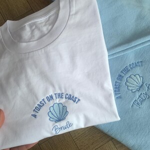 Puede incluir: Camiseta blanca con un dise&ntilde;o de concha bordada en azul y el texto "A Toast on the Coast Bride". En el fondo se ve una sudadera azul claro, tambi&eacute;n con el texto "A Toast on the Coast" y un dise&ntilde;o de concha.