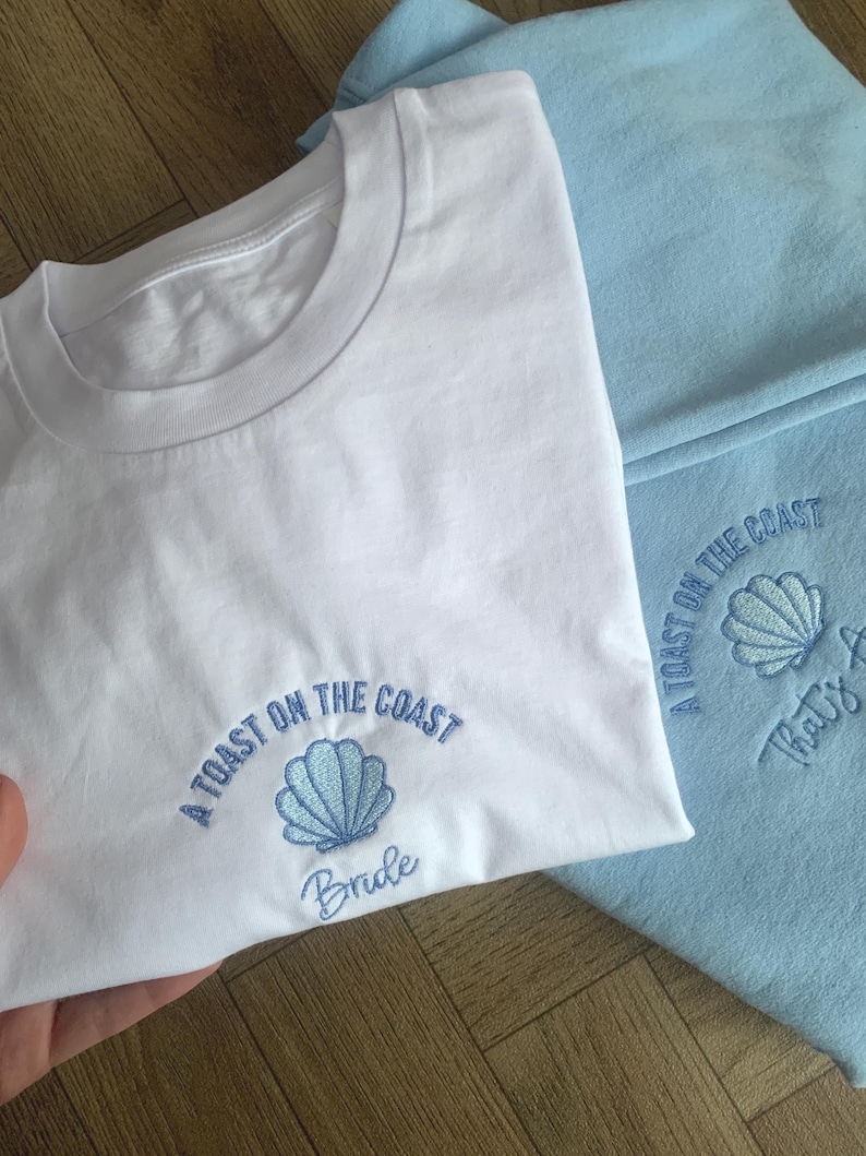Puede incluir: Camiseta blanca con un dise&ntilde;o de concha bordada en azul y el texto "A Toast on the Coast Bride". En el fondo se ve una sudadera azul claro, tambi&eacute;n con el texto "A Toast on the Coast" y un dise&ntilde;o de concha.