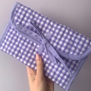 May include: A quilted, lavender and white gingham patterned clutch with a matching bow closure. The clutch has a rounded flap and is held in a hand against a light gray background. The fabric is stitched in a diamond pattern.
