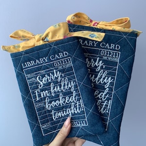May include: Two rectangular dark blue quilted fabric sleeves featuring a white printed library card design with the text "LIBRARY CARD" and "Sorry, I'm fully booked tonight!". Each sleeve is topped with a yellow floral fabric bow.