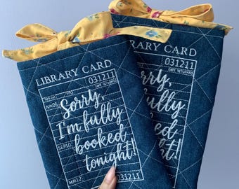 Op de afbeelding: Twee rechthoekige, donkerblauwe, doorgestikte stoffen hoezen met een wit bibliotheekkaartontwerp, inclusief de tekst "LIBRARY CARD" en "Sorry, I'm fully booked tonight!". Elke hoes is afgewerkt met een gele, gebloemde stoffen strik.