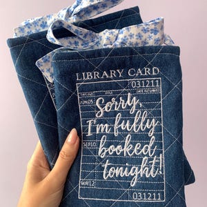 May include: Two quilted denim fabric book sleeves featuring white embroidered library card details and the text Sorry, I'm fully booked tonight! Each sleeve has a blue and white floral patterned fabric tie at the top for closure.