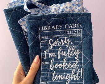 Op de afbeelding: Twee gewatteerde boekenhoezen van denim met wit geborduurde bibliotheekkaartdetails en de tekst Sorry, I'm fully booked tonight! Elke hoes heeft een blauw-witte stoffen strik met bloemenpatroon aan de bovenkant.