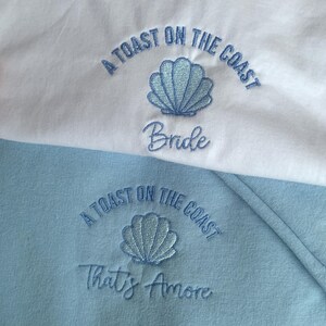 Puede incluir: Dos sudaderas, una blanca y otra azul claro, con texto bordado y dise&ntilde;os de conchas marinas. La sudadera blanca dice "A Toast on the Coast Bride" y la azul dice "A Toast on the Coast That's Amore".