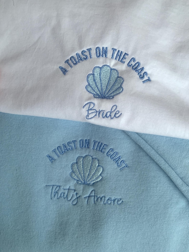 Puede incluir: Dos sudaderas, una blanca y otra azul claro, con texto bordado y dise&ntilde;os de conchas marinas. La sudadera blanca dice "A Toast on the Coast Bride" y la azul dice "A Toast on the Coast That's Amore".