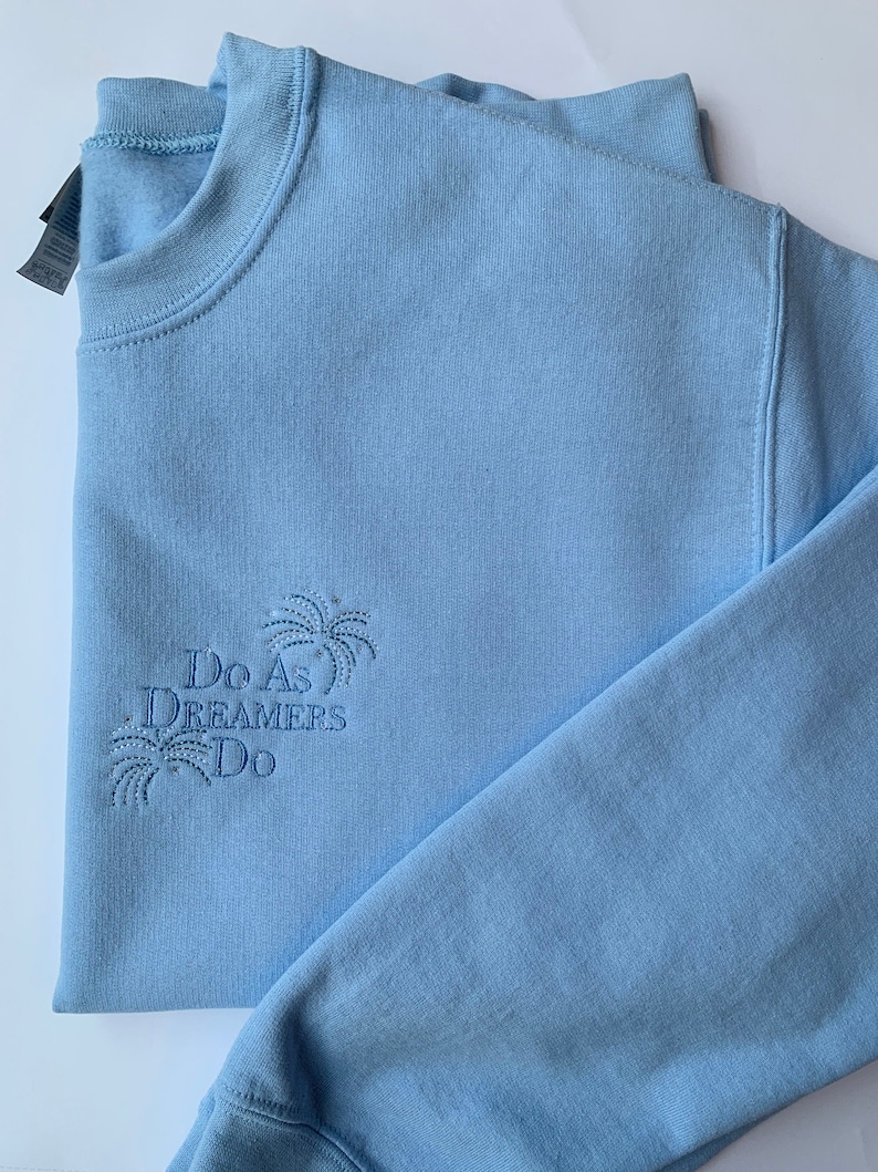 Op de afbeelding: Een lichtblauw sweatshirt met de tekst "Do As Dreamers Do" geborduurd op de voorkant. De tekst wordt omringd door een gestileerd palmboomontwerp.
