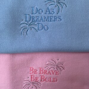 Op de afbeelding: Twee sweatshirts met geborduurde spreuken. De bovenste sweatshirt is lichtblauw met de tekst "Do as Dreamers Do" en een zilveren glitter vuurwerkontwerp. De onderste sweatshirt is roze met de tekst "Be Brave Be Bold" en een zilveren glitter vuurwerkontwerp.