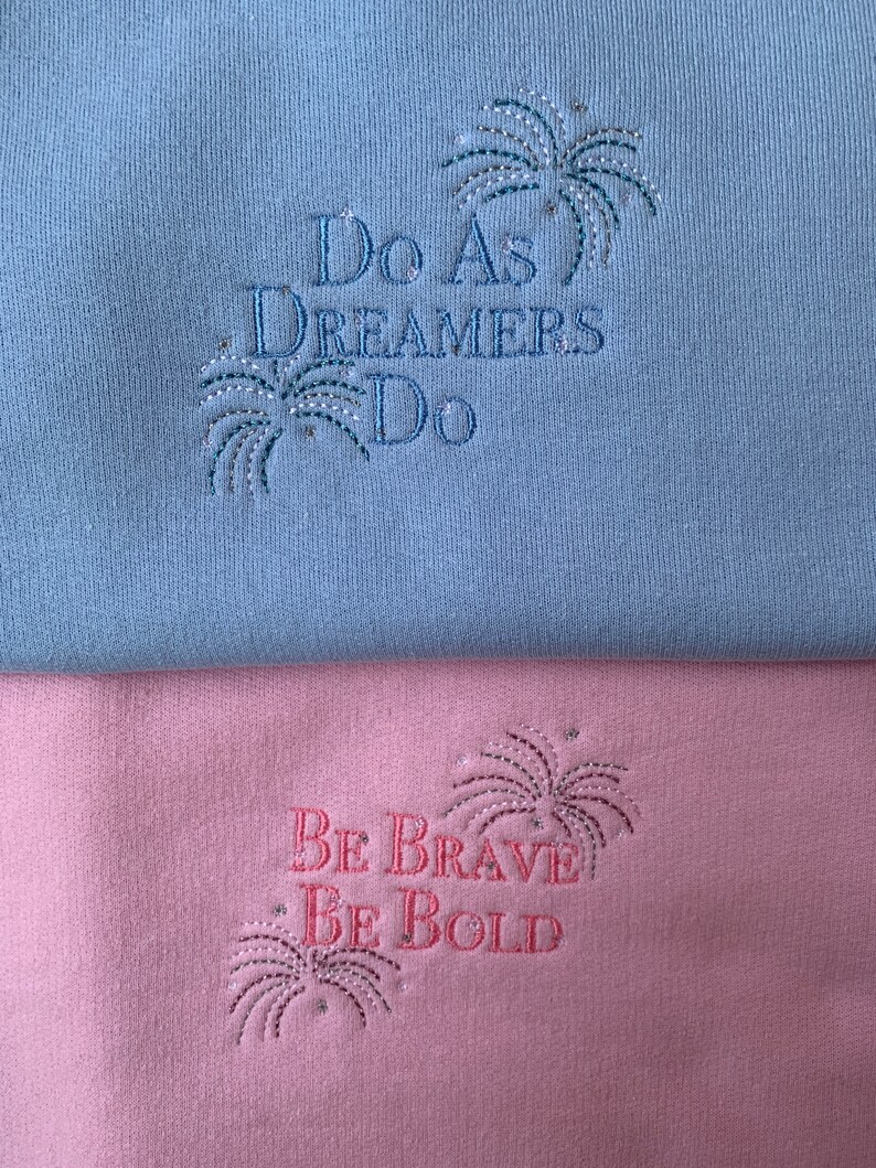 Op de afbeelding: Twee sweatshirts met geborduurde spreuken. De bovenste sweatshirt is lichtblauw met de tekst "Do as Dreamers Do" en een zilveren glitter vuurwerkontwerp. De onderste sweatshirt is roze met de tekst "Be Brave Be Bold" en een zilveren glitter vuurwerkontwerp.