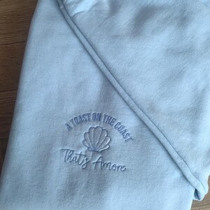 Puede incluir: Sudadera azul claro con texto bordado y dise&ntilde;o de concha. El texto dice "A Toast on the Coast" encima de "That's Amore". La sudadera est&aacute; doblada, mostrando la manga y la capucha.