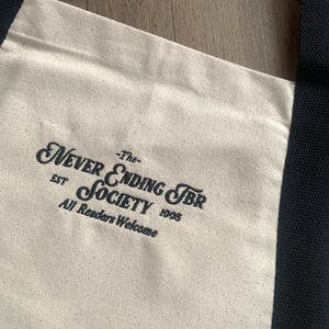 May include: Cream-colored tote bag with black handles and trim. The bag features the text "Never Ending TBR Society" in a decorative font, with "EST 1995" and "All Readers Welcome" below. The bag is made of a canvas-like material.