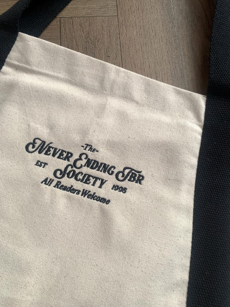 May include: Cream-colored tote bag with black handles and trim. The bag features the text "Never Ending TBR Society" in a decorative font, with "EST 1995" and "All Readers Welcome" below. The bag is made of a canvas-like material.