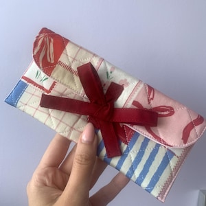 May include: A quilted clutch with a patchwork design featuring red, pink, and blue patterns. The clutch is secured with a dark red bow. The design includes floral and striped patterns.