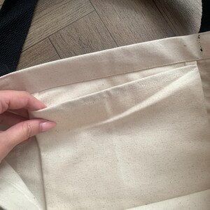 May include: Cream-colored canvas tote bag with a black strap. The interior features a pocket. The bag is open, revealing the inside. The background is a light brown wooden surface.