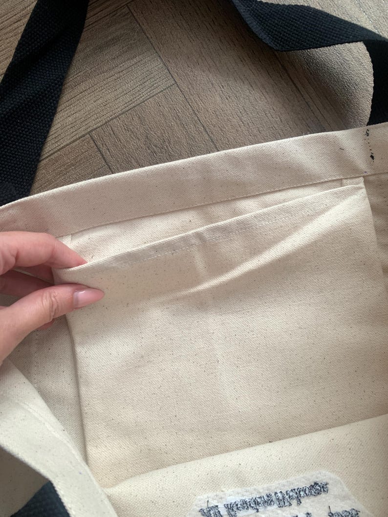 May include: Cream-colored canvas tote bag with a black strap. The interior features a pocket. The bag is open, revealing the inside. The background is a light brown wooden surface.