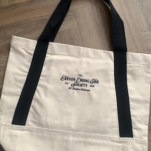 May include: Cream-colored tote bag with black handles. The bag features the text "The Never Ending TBR Society" in black script, with "EST 1995" and "All Readers Welcome" below. The bag is made of a sturdy canvas material.