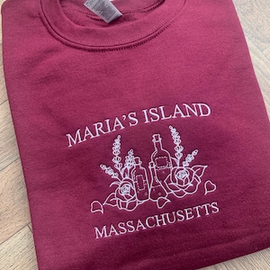 May include: A maroon sweatshirt featuring white embroidered text and a floral design. The text reads "MARIA'S ISLAND MASSACHUSETTS". The design includes bottles, flowers, and sprigs.