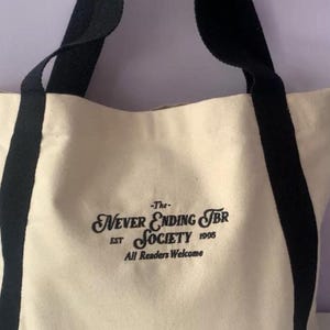 May include: Cream-colored tote bag with black straps and accents. The bag features the text "-The- Never Ending TBR Society EST 1995 All Readers Welcome" in a decorative font. The bag is made of a sturdy canvas material.
