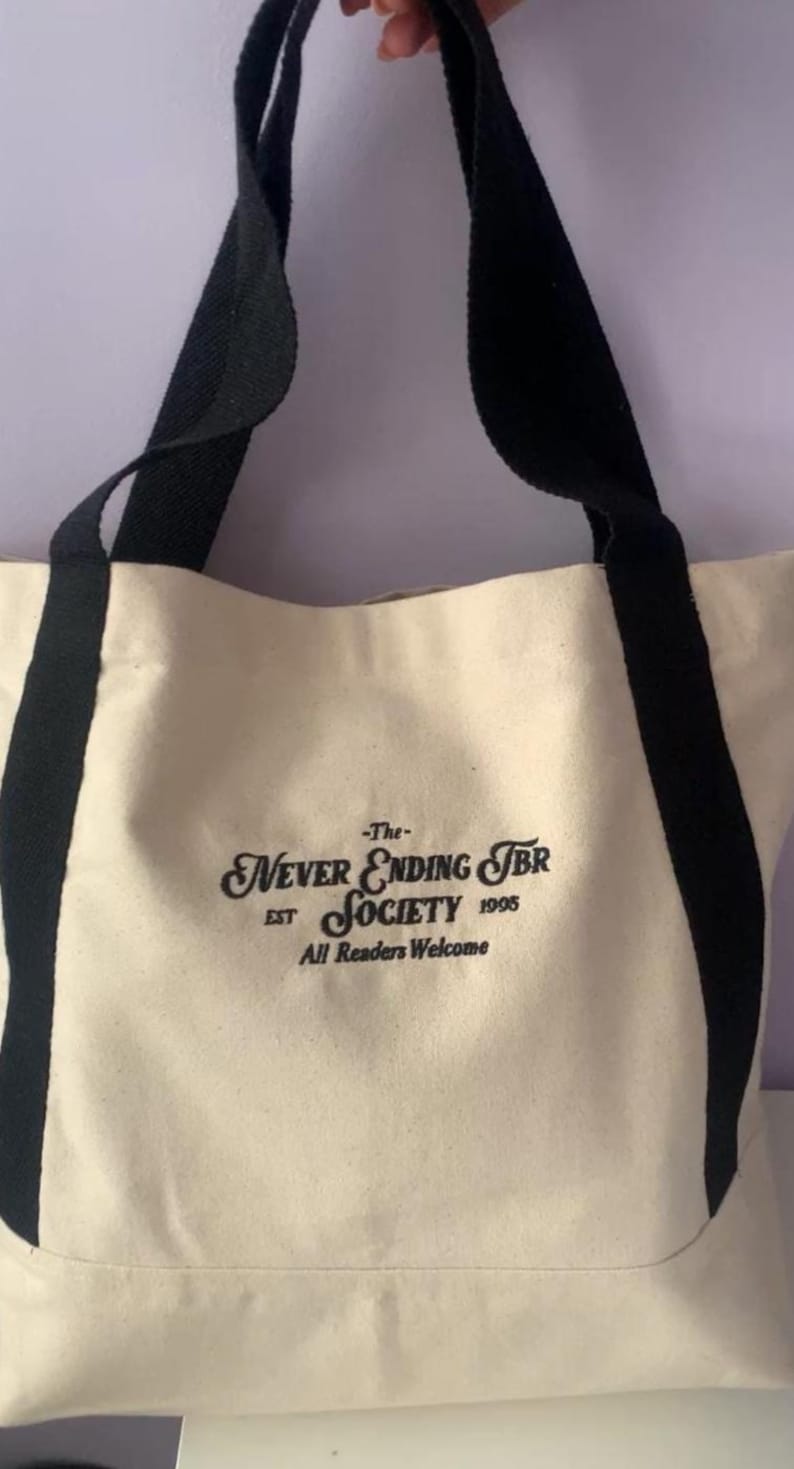 May include: Cream-colored tote bag with black straps and accents. The bag features the text "-The- Never Ending TBR Society EST 1995 All Readers Welcome" in a decorative font. The bag is made of a sturdy canvas material.