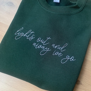 May include: A dark green sweatshirt with the text "lights out and away we go" embroidered in white.