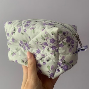 May include: A quilted cosmetic bag with a floral pattern. The bag is white with a repeating pattern of purple flowers and green leaves. It has a silver zipper closure and a small fabric loop on the side.
