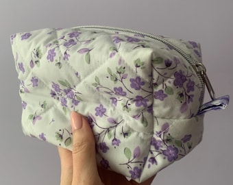 Op de afbeelding: Een gewatteerde make-uptas met een bloemenpatroon. De tas is wit met een herhalend patroon van paarse bloemen en groene bladeren. Het heeft een zilveren ritssluiting en een kleine stoffen lus.