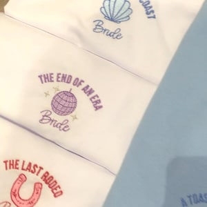 Puede incluir: Telas blancas con texto y gr&aacute;ficos bordados. Una dice "A Toast on the Coast Bride" con una concha. Otra dice "The End of an Era Bride" con una bola de discoteca. La &uacute;ltima dice "The Last Rodeo Bride" con una herradura.