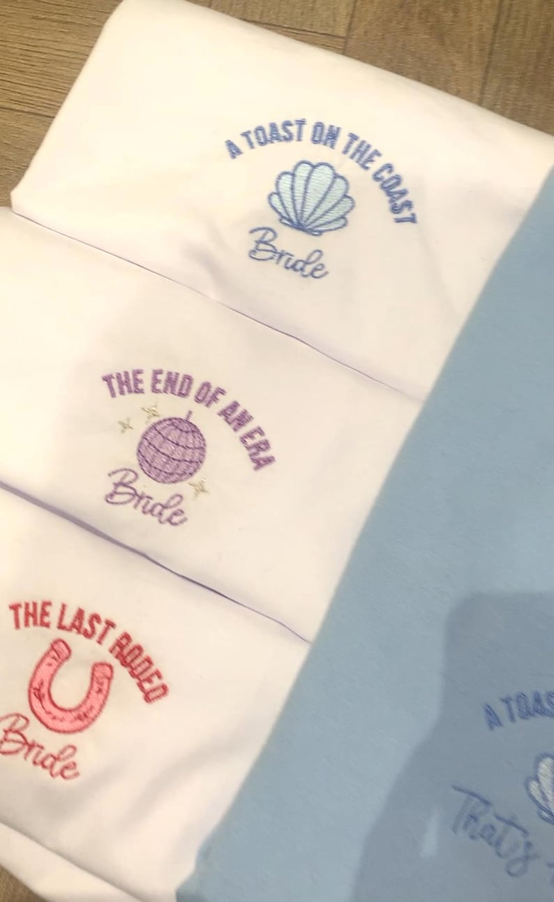Puede incluir: Telas blancas con texto y gr&aacute;ficos bordados. Una dice "A Toast on the Coast Bride" con una concha. Otra dice "The End of an Era Bride" con una bola de discoteca. La &uacute;ltima dice "The Last Rodeo Bride" con una herradura.