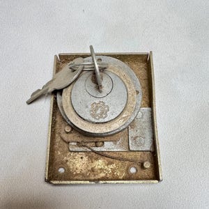 May include: A vintage metal door lock with a key attached. The lock is rectangular and has a circular keyhole. The lock is rusty and has a worn finish.