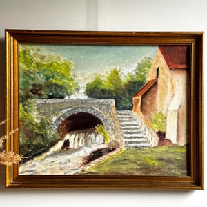 Puede incluir: Pintura al óleo en un marco dorado que representa un puente de piedra sobre una cascada, con un edificio y escaleras a la derecha. La obra presenta árboles verdes y un cielo nublado, con la firma del artista en la esquina inferior derecha.