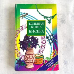 Op de afbeelding: Een boek getiteld "БОЛЬШАЯ КНИГА БИСЕРА" (Groot Boek van Kralen) met een groene, gele en paarse omslag. De omslag toont kralenkunst, waaronder een vaas met bloemen, sieraden en een kleine tas. Het boek is een gids voor kralenwerk.