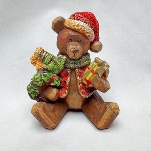 May include: A brown bear figurine wearing a red Santa hat with gold trim and a green and red scarf. The bear holds a wrapped gift with a yellow bow. The figurine is a festive holiday decoration.