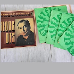 Peut inclure: Un coffret de disques vinyles vintage avec un portrait de Georg Ots, un chanteur soviétique. La pochette de l'album est brune avec des lettres dorées et le texte "РОДНЫЙ АРТИСТ СССР ГЕОРГ ОЦС SV LIIDU RAHVAKUNSTNIK" est visible. Les pochettes de disques sont vertes avec un motif répété de violons stylisés.