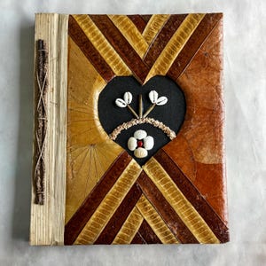 Puede incluir: Un diario hecho a mano con un recorte en forma de corazón, decorado con conchas marinas y un patrón de hojas. La cubierta presenta una mezcla de tonos marrones y beige, con una encuadernación con cuerda.
