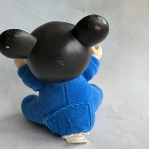 Op de afbeelding: Een blauw pluche Mickey Mouse speelgoed dat zit met gekruiste armen. Het speelgoed heeft zwarte oren en een zwart hoofd. Het speelgoed draagt een blauwe jumpsuit.