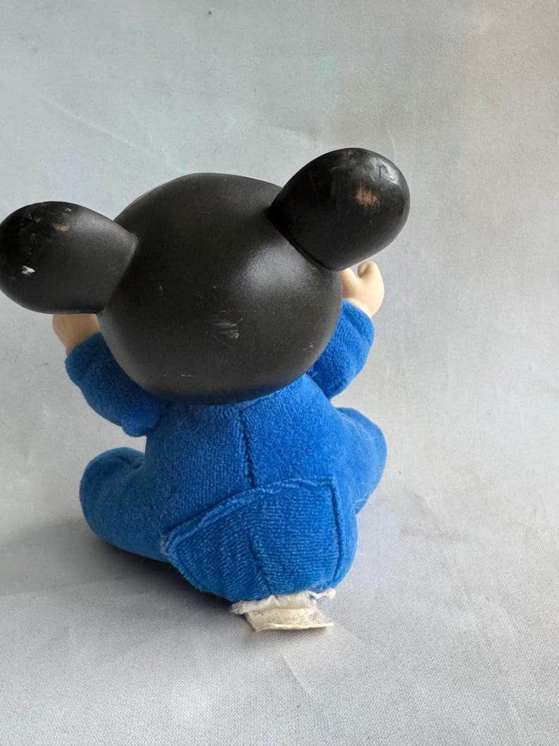 Op de afbeelding: Een blauw pluche Mickey Mouse speelgoed dat zit met gekruiste armen. Het speelgoed heeft zwarte oren en een zwart hoofd. Het speelgoed draagt een blauwe jumpsuit.