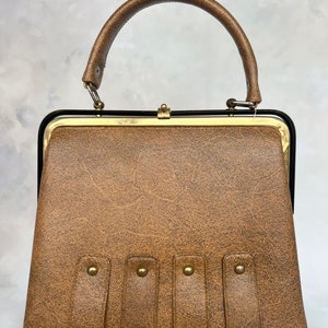 Könnte beinhalten: Eine braune Ledertasche mit goldenem Rahmen und einem oberen Henkel. Die Tasche hat eine strukturierte Oberfläche und vier goldene Nieten auf der Vorderseite.