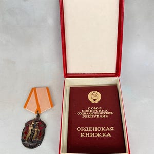 May include: A vintage medal with a red and orange ribbon, displayed in an open red box. The medal features a detailed design with figures and text. A maroon booklet with gold lettering is also in the box.