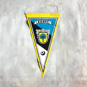 May include: A yellow, black, and white pennant flag with the word "EESTI" in blue and white. The flag features a coat of arms with a blue flower and a white string for hanging.