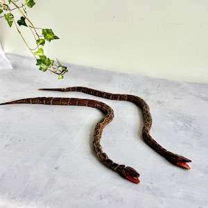 Puede incluir: Dos figuras de serpiente de madera articuladas con cuerpos de color marrón oscuro, intrincados patrones geométricos de color tostado y bocas pintadas de rojo brillante. Las serpientes están segmentadas para permitir un movimiento flexible y se muestran en una posición curva.