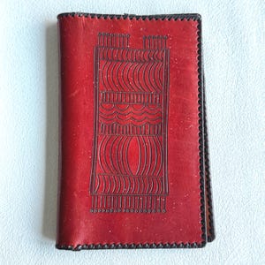 May include: A red leather journal with a decorative, embossed design. The cover features a rectangular pattern with curved and straight lines, and small dots. The edges are stitched with black thread, creating a border.