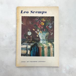 Op de afbeelding: Vintage boekomslag met de titel "Leo Svemps" in blauwe tekst. De omslag toont een schilderij van een bloemstuk in een blauw-witte vaas, een klok en een gestreepte tafelkleed. De naam van de uitgever staat onderaan.