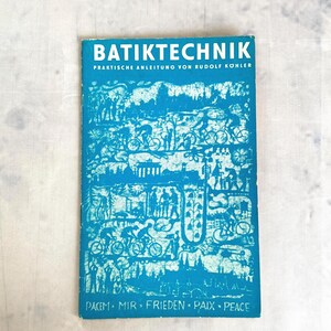Könnte beinhalten: Ein Vintage-Buch mit dem Titel "BATIKTECHNIK" mit blauem Einband und weißen Illustrationen von Radfahrern und Figuren. Das Buch enthält deutschen Text und das Wort "Frieden" in mehreren Sprachen.