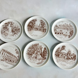 May include: Six white ceramic coasters with brown illustrations of buildings and landmarks. Each coaster features the text "Bad Pyrmont" and the name of the landmark.