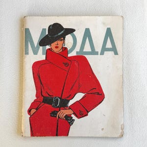 May include: Vintage magazine cover featuring a fashion illustration. The illustration depicts a woman in a red coat, black hat, and gloves. The word "MOAA" is printed in light blue letters at the top of the cover.
