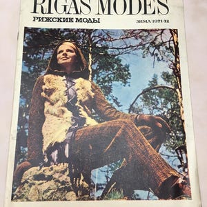 Può includere: Una copertina di rivista con una donna che indossa una giacca marrone con bordatura in pelliccia e pantaloni marroni. La rivista è intitolata "Rīgas Modes" con il sottotitolo "Rīdzские Моды" e la data "зима 1971-72".