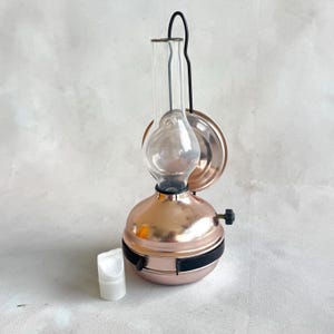 May include: A copper-colored miniature oil lamp with a clear glass chimney and a black handle. The lamp has a round base and a small, white, cylindrical container next to it. The lamp is a decorative item.