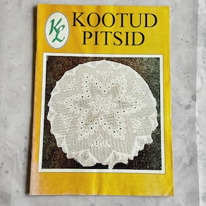 Puede incluir: Un libro vintage con una cubierta amarilla con el texto "KOOTUD PITSID" y un tapete de encaje blanco. El tapete tiene un diseño floral detallado. El libro tiene un logotipo verde y blanco en la esquina superior izquierda.