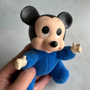 Op de afbeelding: Een blauw pluche Mickey Mouse speelgoed met grote ogen en een zwarte neus. Het speelgoed draagt een blauwe romper.