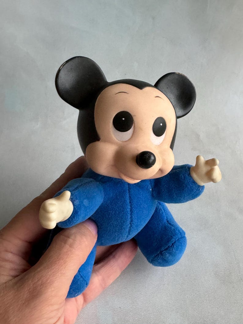 Op de afbeelding: Een blauw pluche Mickey Mouse speelgoed met grote ogen en een zwarte neus. Het speelgoed draagt een blauwe romper.